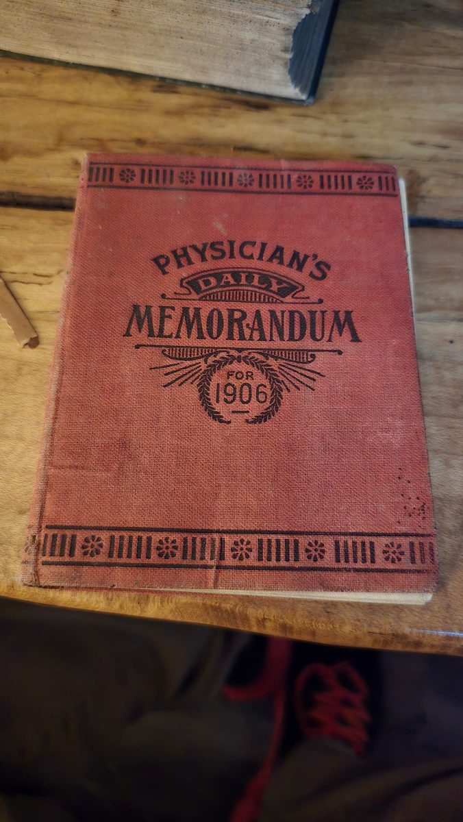 Vintage 1906 Physician’s Daily Memorandum in well-worn red cloth cover, displayed on wooden table.