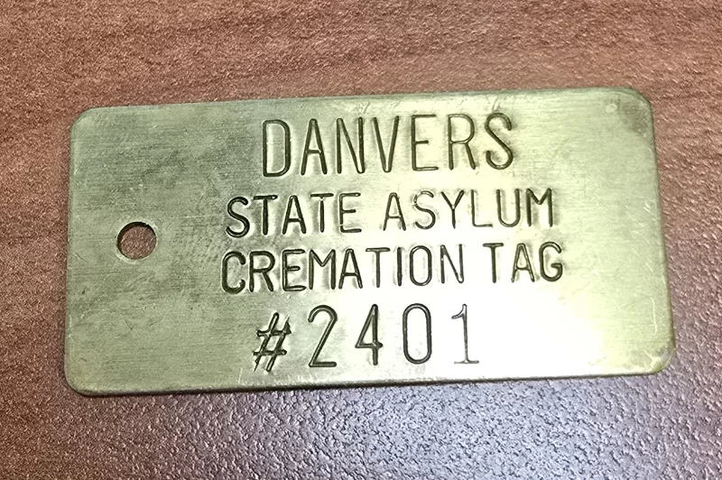 Brass cremation tag replica labeled Danvers State Asylum Cremation Tag #2401, engraved and aged.