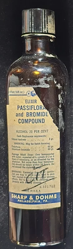 Image of a vintage brown glass bottle labeled “Elixir Passiflora and Bromide Compound,” manufactured by Sharp & Dohme, Philadelphia, PA. The label indicates a 20% alcohol content and includes ingredients such as chloral hydrate, passiflora, and strontium bromide. Label shows signs of wear and partial tearing.