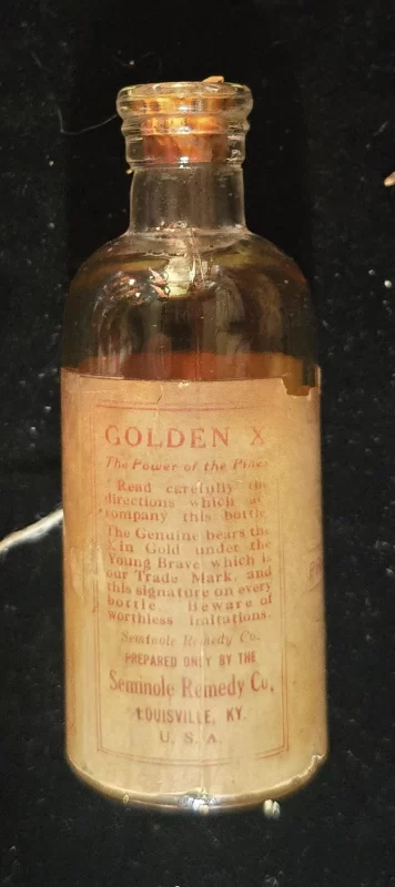 Front view of antique “Golden X” bottle with amber fluid, cork stopper, and aged label reading “Seminole Remedy Co., Louisville, KY.” Partial directions and promotional text visible.