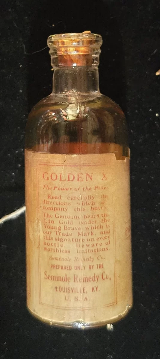 Front view of antique “Golden X” bottle with amber fluid, cork stopper, and aged label reading “Seminole Remedy Co., Louisville, KY.” Partial directions and promotional text visible.