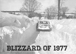 Car driving between towering snowbanks after the Blizzard of 1977, with snow piled high on both sides of a narrow rural road.