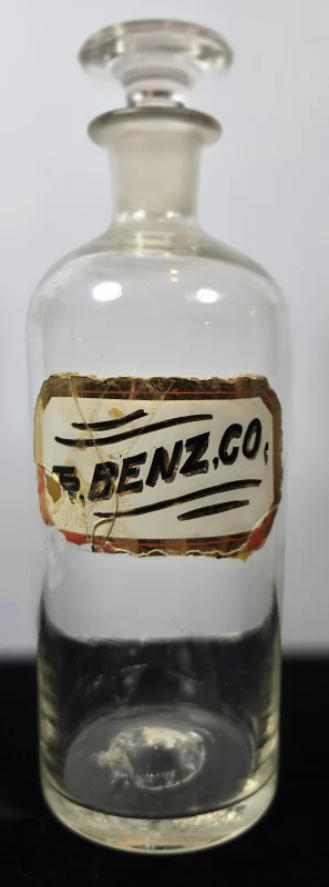 Clear glass apothecary bottle labeled T. Benz. Co. with ground glass stopper.