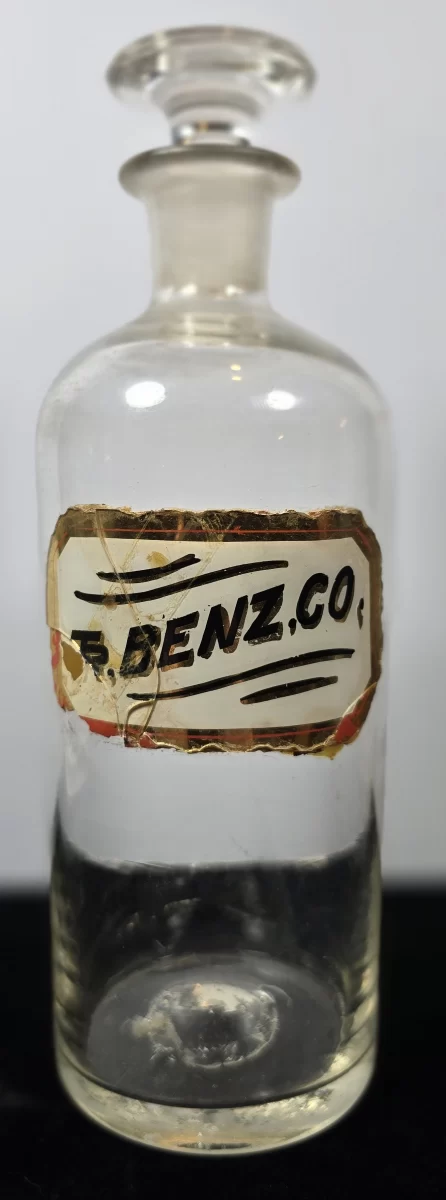 Clear glass apothecary bottle labeled T. Benz. Co. with ground glass stopper.