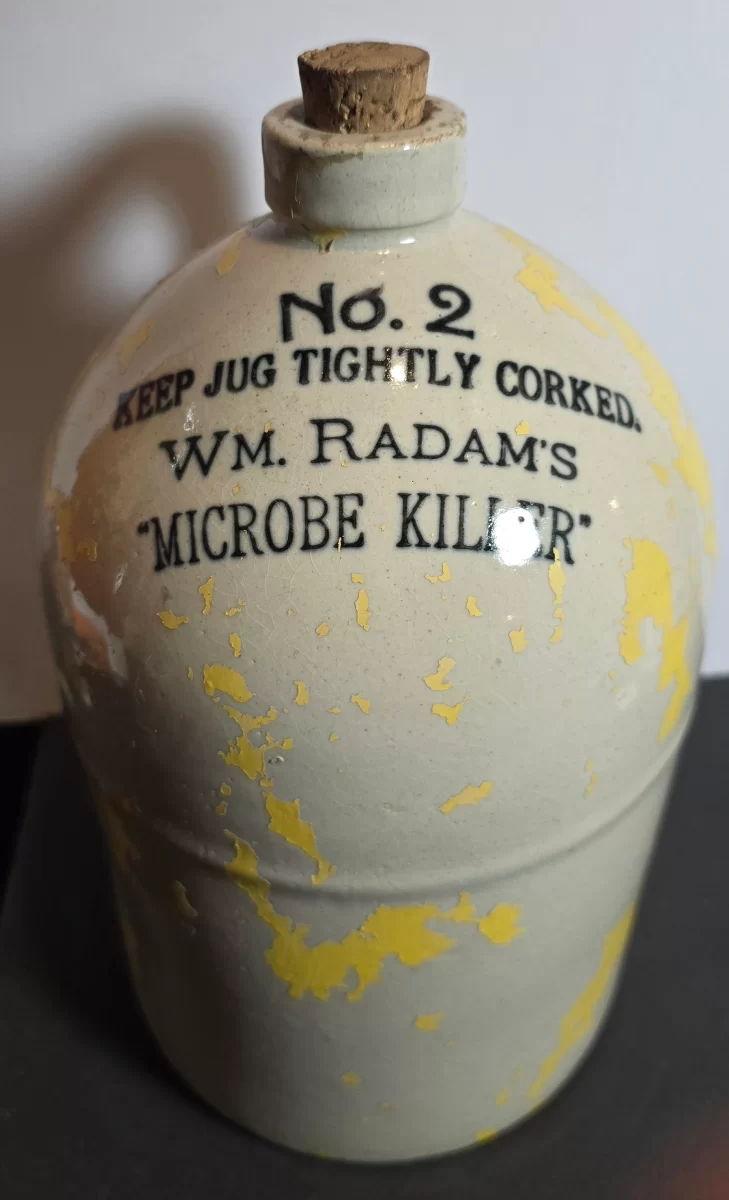 Stoneware jug marked “Wm. Radam’s Microbe Killer No. 2” with aged glaze, yellow paint loss, and cork stopper.