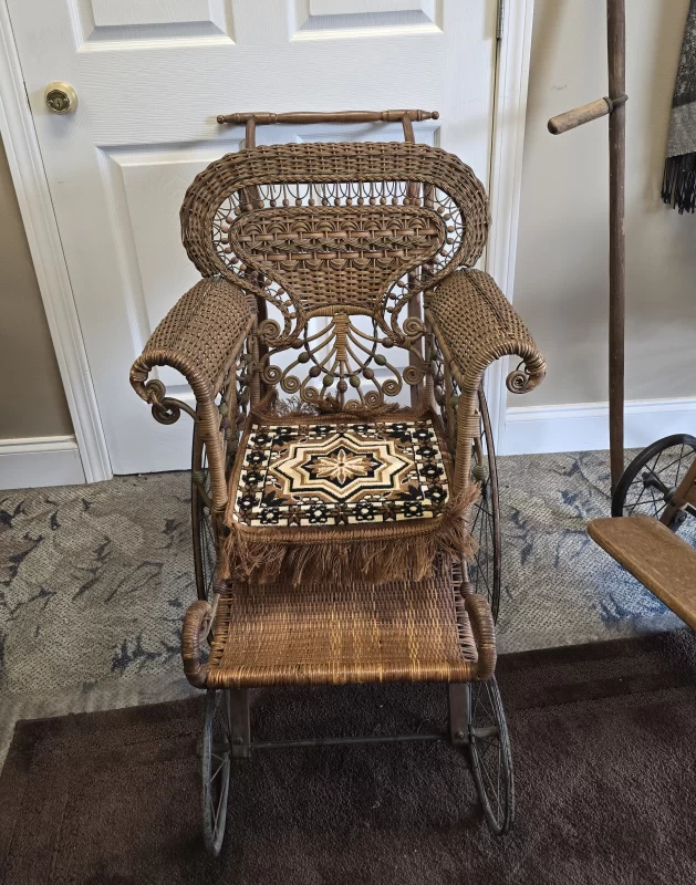 Victorian pediatric wicker invalid chair with ornate scrollwork, spoked wheels, and child-sized seat.
