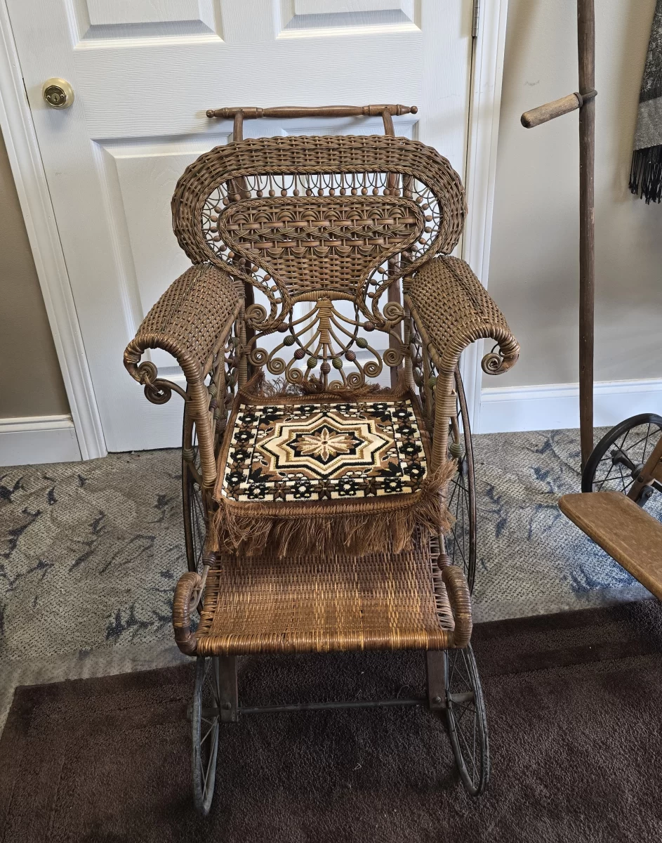 Victorian pediatric wicker invalid chair with ornate scrollwork, spoked wheels, and child-sized seat.